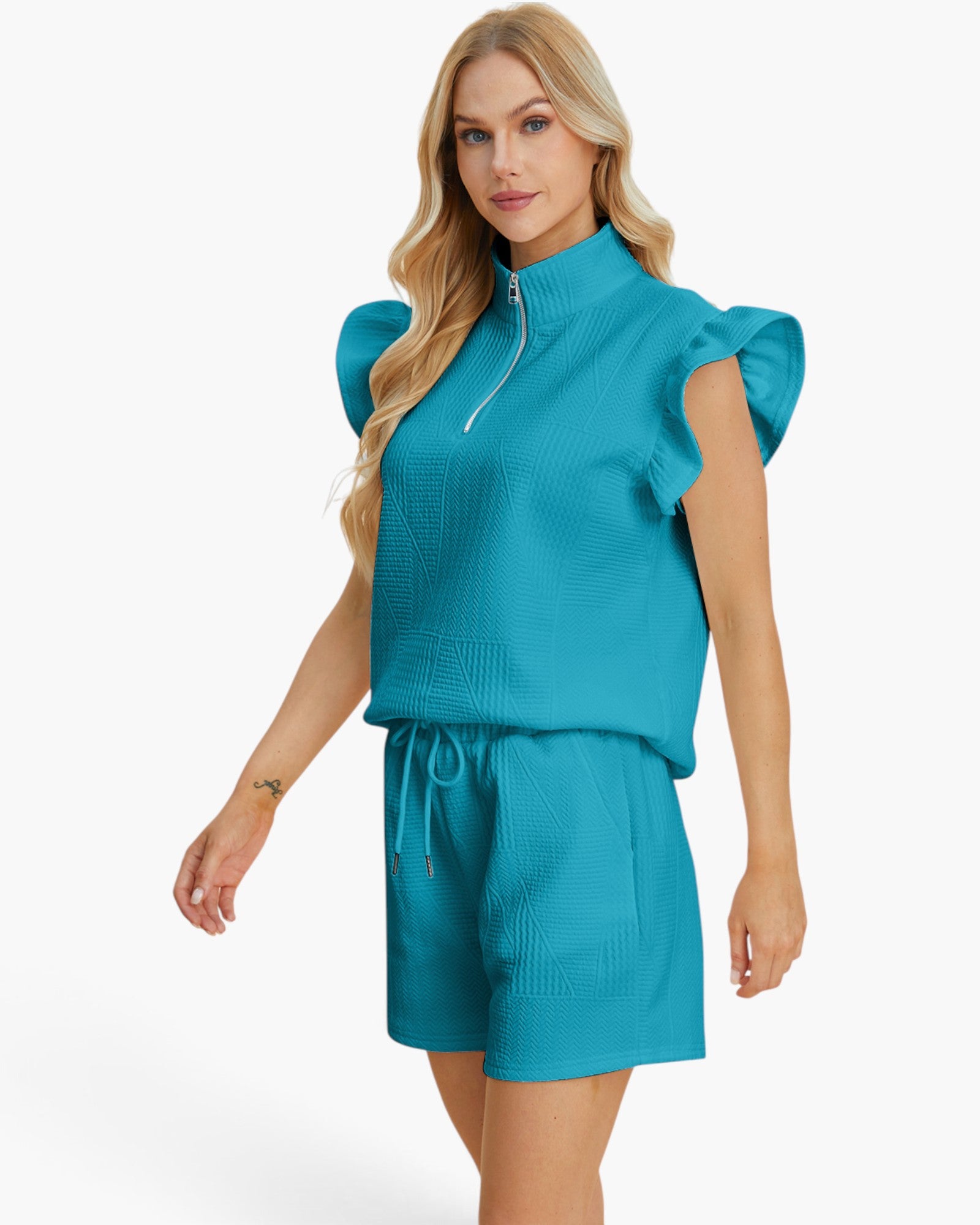 A blonde model wearing a matching turquoise blue two-piece set consisting of a textured ruffle-sleeve mock-neck top with a half-zip and matching drawstring shorts, set against a plain white studio background.