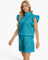 A blonde model wearing a matching turquoise blue two-piece set consisting of a textured ruffle-sleeve mock-neck top with a half-zip and matching drawstring shorts, set against a plain white studio background.