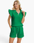 A woman modeling a bright green two-piece lounge set featuring a short-sleeve top with ruffled sleeves and a half-zip collar, paired with matching drawstring shorts, against a light gray studio background.