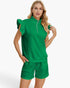 A woman modeling a bright green two-piece lounge set featuring a short-sleeve top with ruffled shoulders and a half-zip neckline, paired with matching textured shorts, against a light gray studio background.