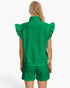 A model shown from the back wearing a bright green textured knit matching set consisting of a sleeveless mock-neck top with ruffled shoulders and matching shorts, against a plain white studio background.