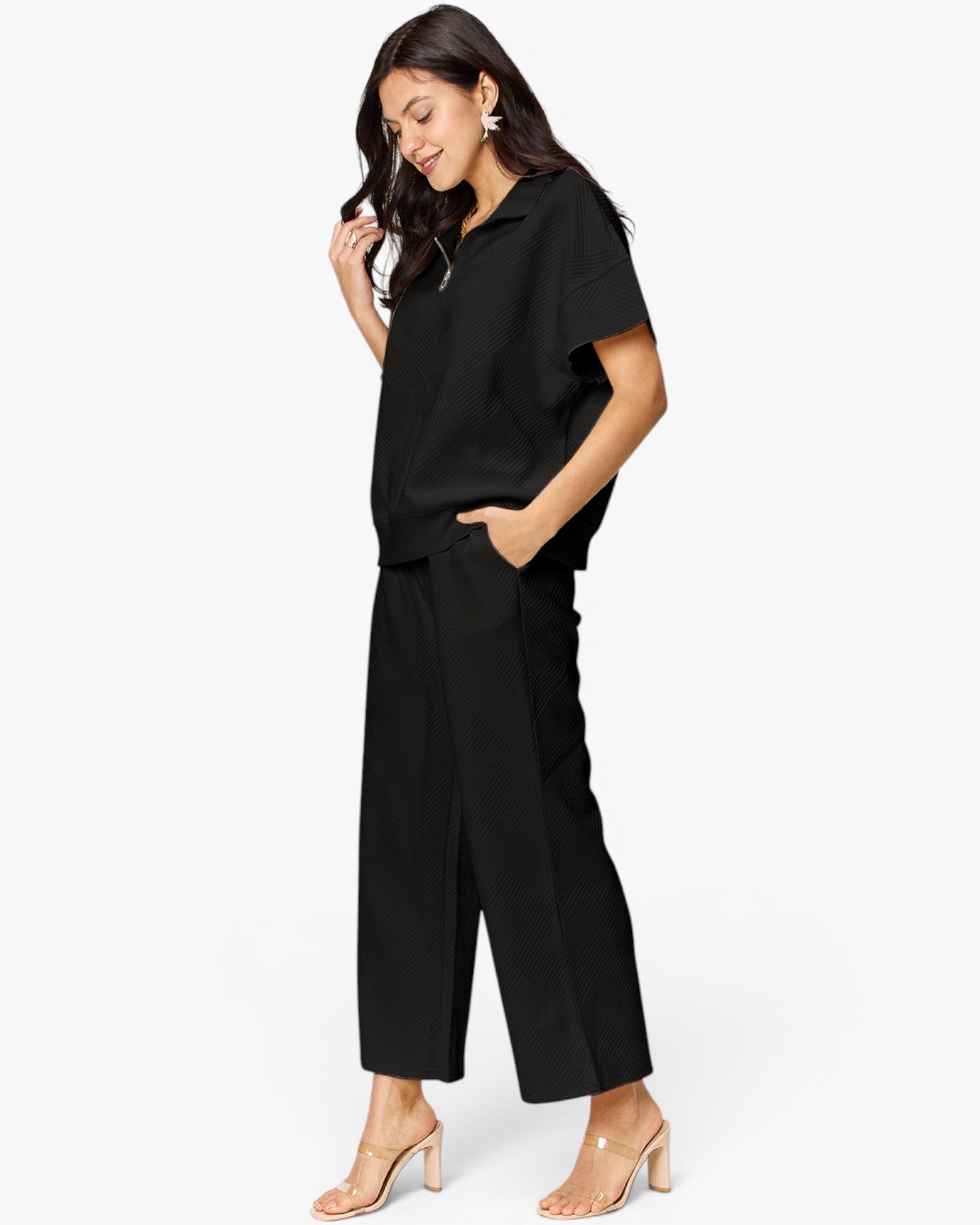 A woman modeling a black short-sleeve collared top and matching black wide-leg cropped pants, paired with clear-strap heeled sandals, against a plain white studio background.