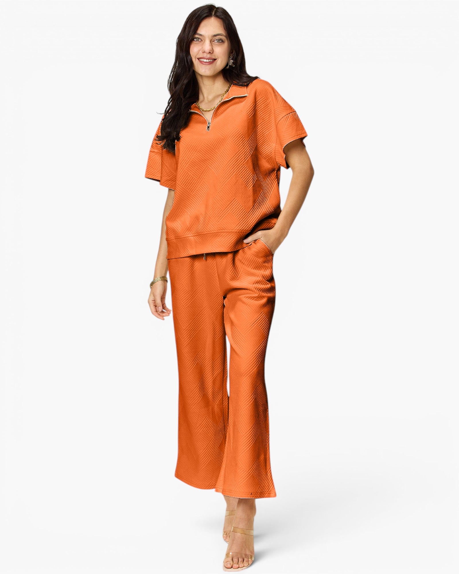 A woman modeling a matching two-piece lounge set in a vibrant orange color, featuring a short-sleeve half-zip top and wide-leg pants, set against a plain white studio background.