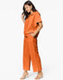A woman modeling a matching burnt orange short-sleeve collared top and wide-leg cropped pants set, paired with clear strap high-heeled sandals, against a plain white studio background.