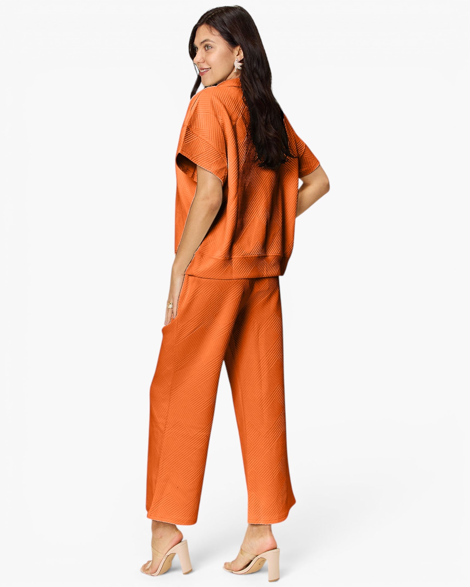 A woman modeling a matching two-piece orange outfit consisting of a short-sleeve cropped top and wide-leg cropped pants, shown from a three-quarters back view against a plain white studio background.