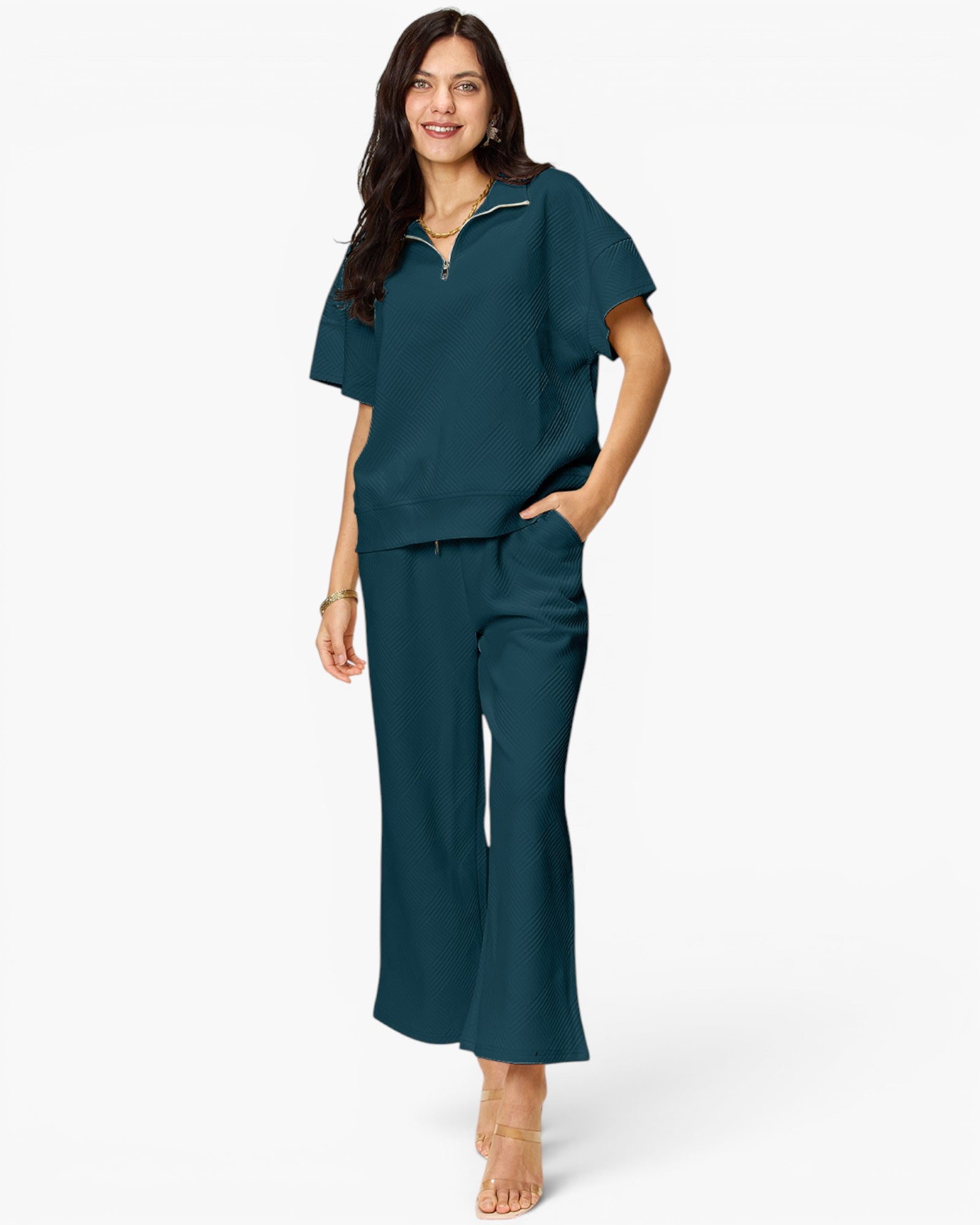 A woman modeling a matching two-piece scrub set in a deep teal color, featuring a short-sleeve half-zip top and matching wide-leg pants, set against a plain white studio background.