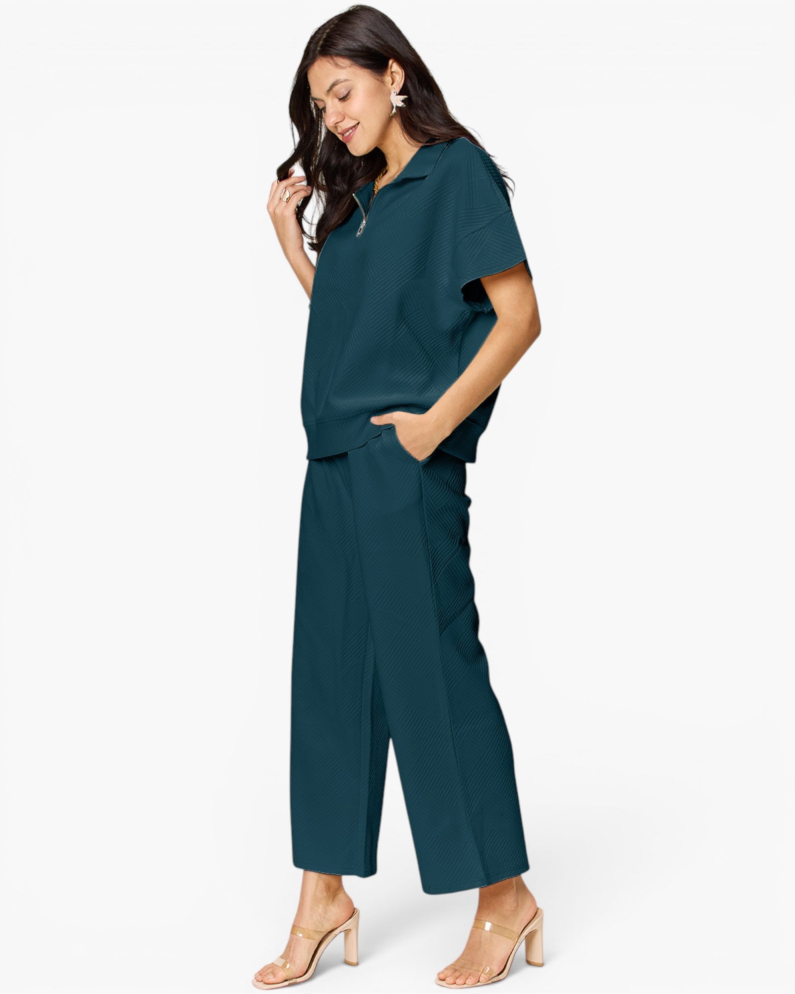 A woman modeling a dark teal two-piece set consisting of a short-sleeve collared top and matching wide-leg cropped pants, paired with nude heeled sandals against a plain white studio background.