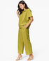 A woman modeling a matching olive green two-piece set consisting of a short-sleeve collared top and wide-leg cropped pants, paired with clear high-heeled sandals against a plain white background.