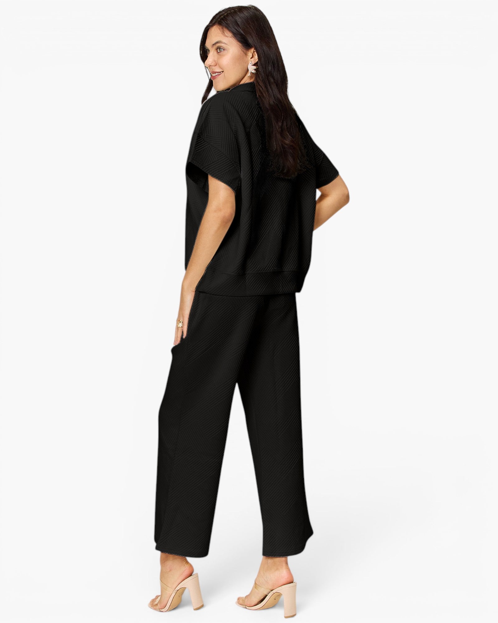 A woman modeling a black short-sleeve top and matching wide-leg cropped pants, shown from a side-back angle against a plain light gray studio background.