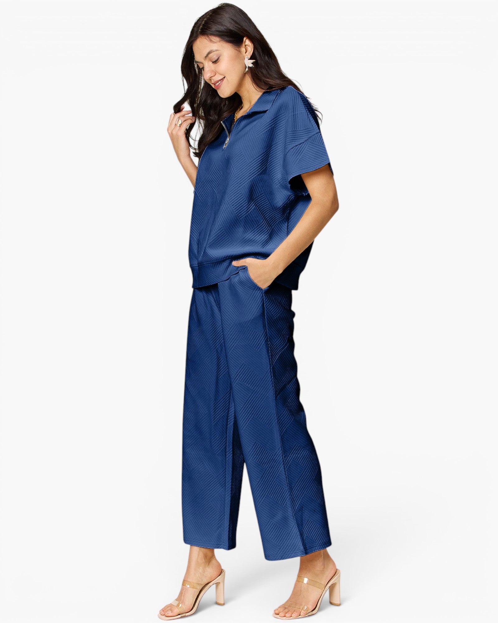 A woman modeling a matching navy blue two-piece set consisting of a short-sleeve collared top and wide-leg cropped pants, paired with clear-strap heeled sandals against a plain white studio background.
