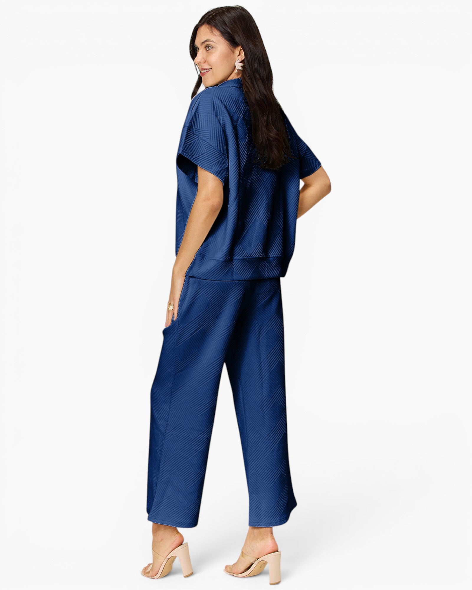 A woman modeling a matching navy blue two-piece set consisting of a short-sleeve cropped top and wide-leg cropped pants, shown from a three-quarters back view against a plain white studio background.