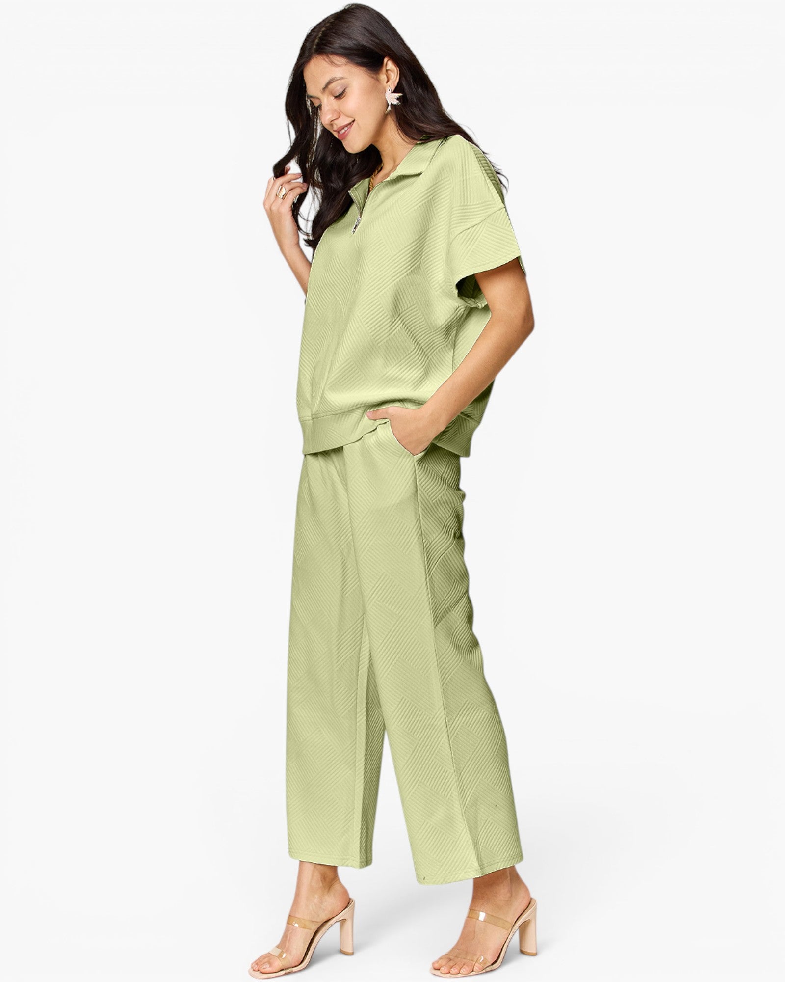 A woman modeling a light sage green matching set consisting of a short-sleeve collared top and wide-leg cropped pants, paired with clear-strap heeled sandals against a plain white studio background.