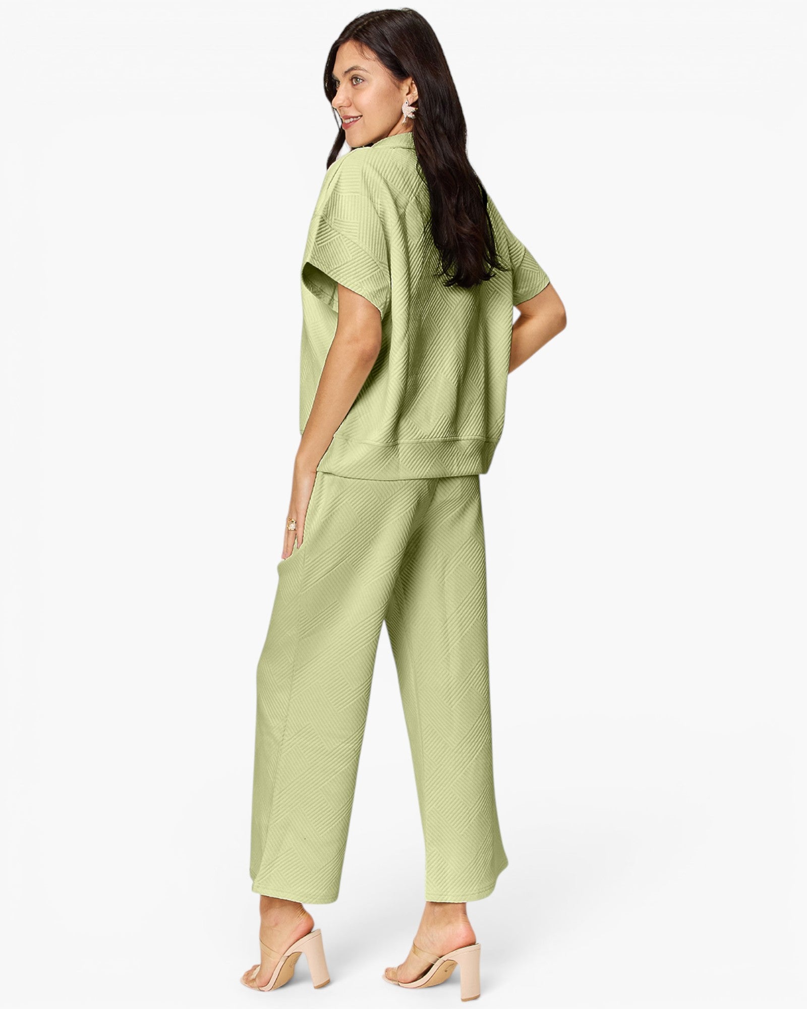 A woman modeling a light sage green two-piece set consisting of a short-sleeve cropped top and matching wide-leg cropped pants, shown from a three-quarters back view against a plain white studio background.