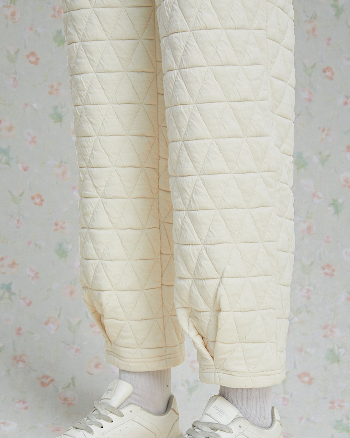 Quilted cream-colored pants worn with white sneakers on a light floral-patterned background