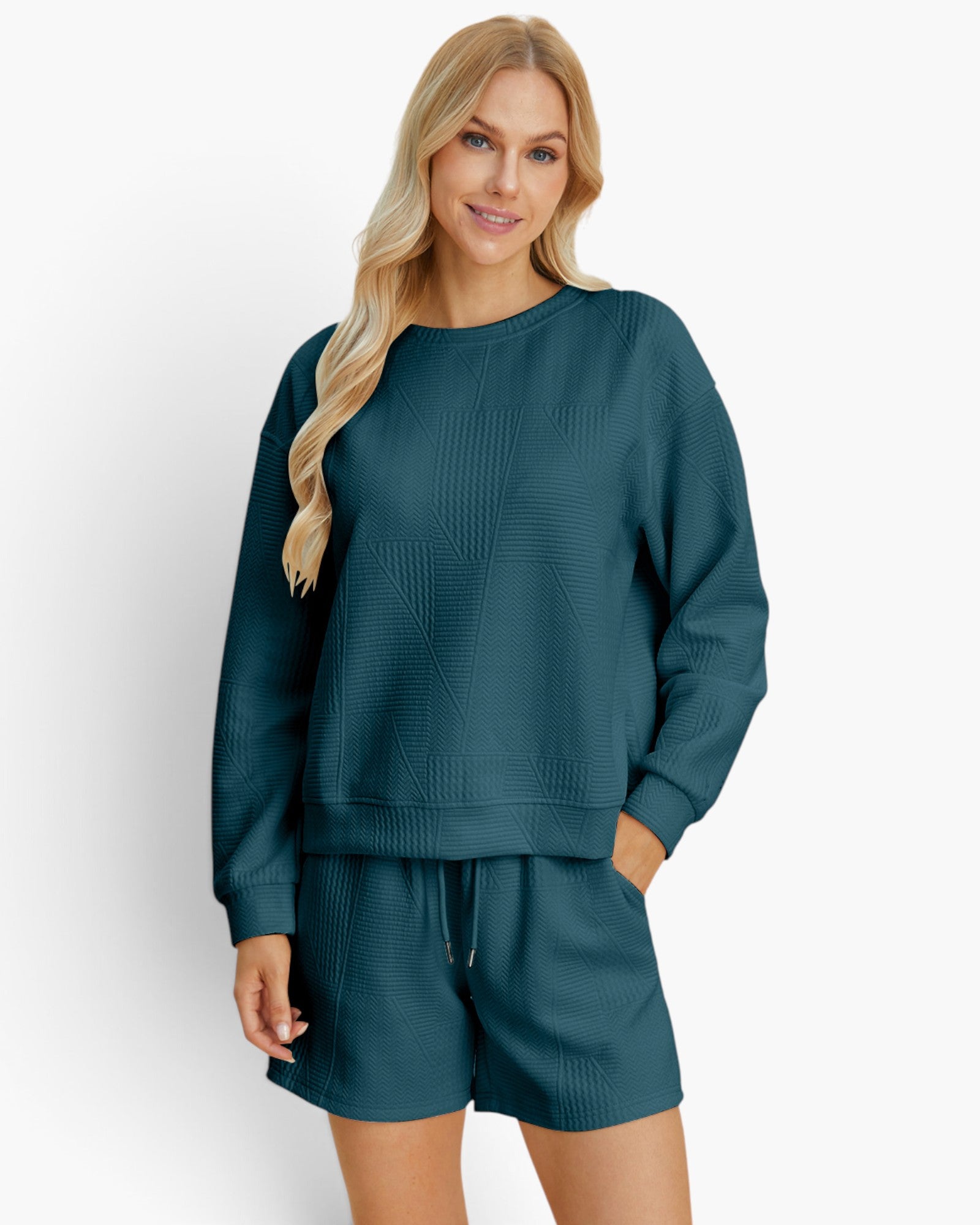A woman modeling a matching dark teal green lounge set consisting of a textured long-sleeve sweatshirt and coordinating shorts against a clean white studio background.