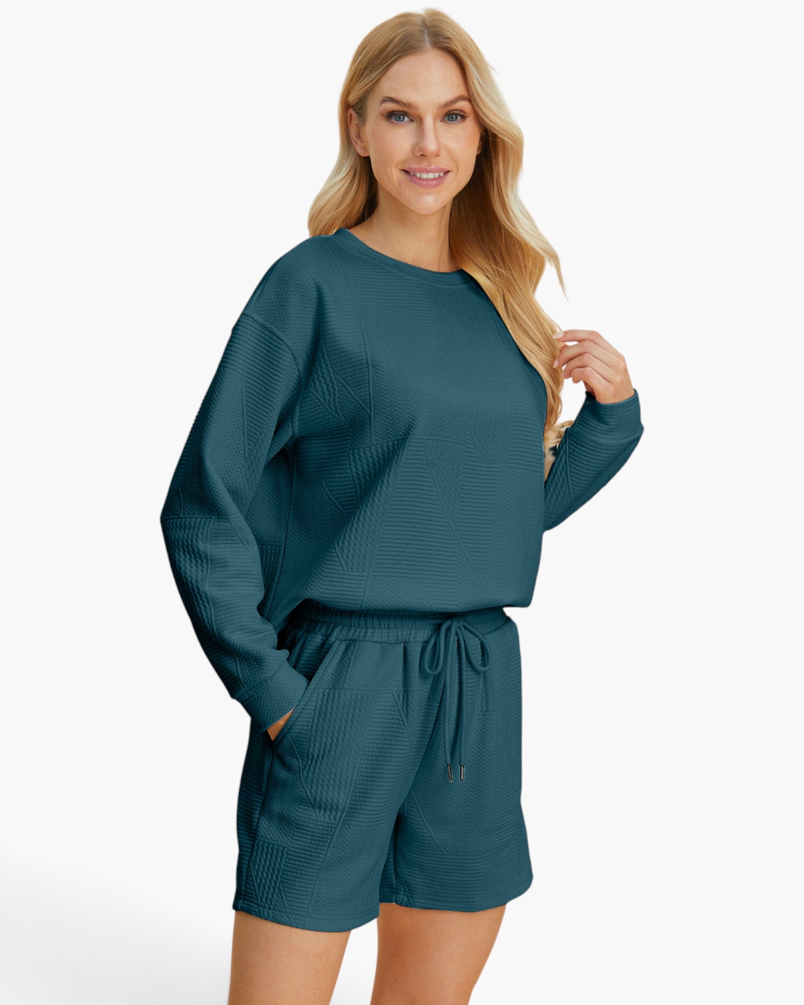 A woman modeling a matching two-piece lounge set consisting of a textured long-sleeve top and drawstring shorts in a deep teal blue-green color, set against a plain light gray background.