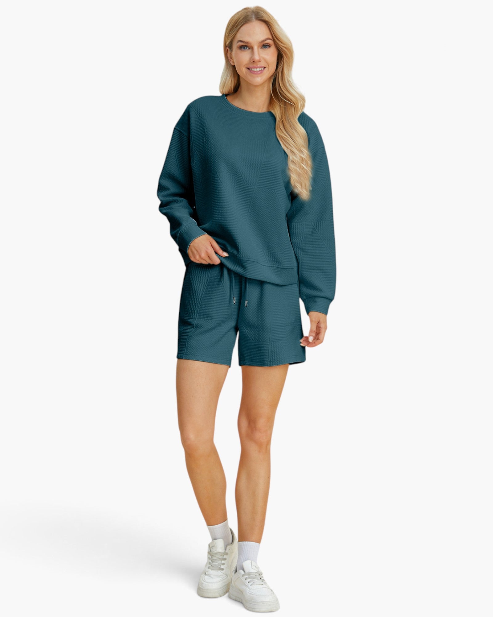 A woman modeling a matching two-piece lounge set consisting of a long-sleeve sweatshirt and shorts in a muted teal blue-green color, paired with white sneakers and socks, against a light gray studio background.