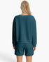 A woman viewed from the back wearing a dark teal textured long-sleeve sweatshirt and matching shorts against a clean white studio background.
