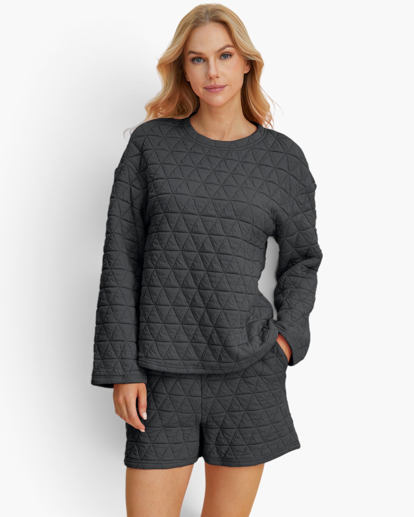 A woman modeling a dark gray quilted matching lounge set consisting of a long-sleeve top and shorts, against a plain white studio background.