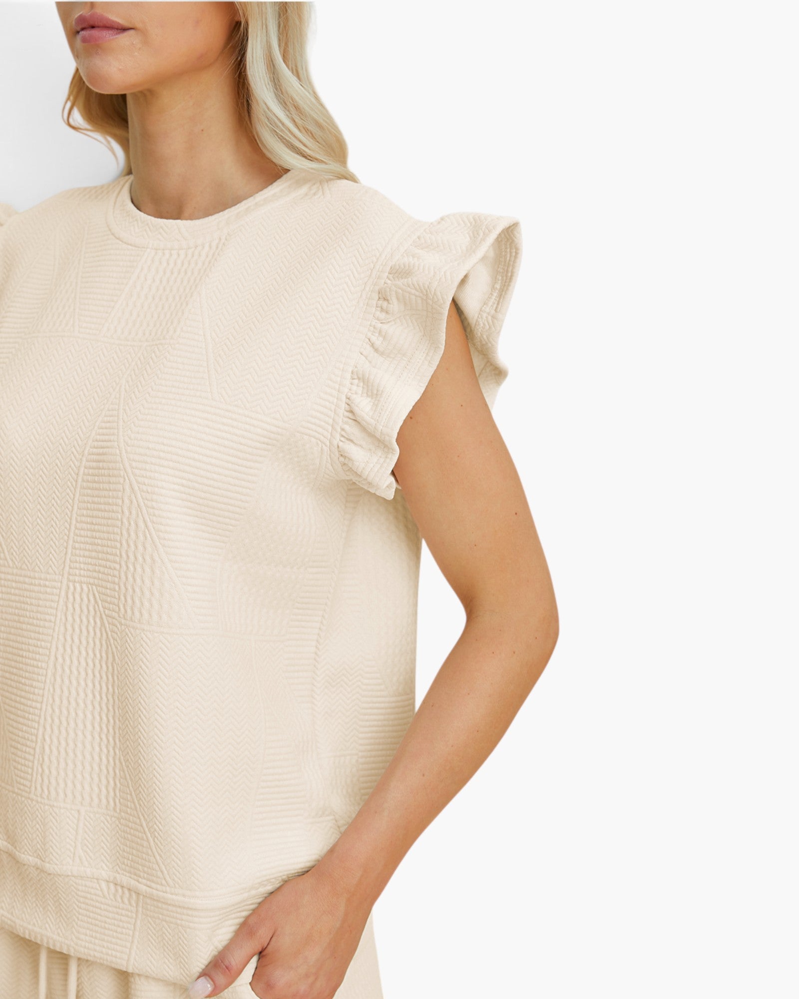 A close-up of a model wearing a cream-colored sleeveless top with textured vertical panels and ruffled cap sleeves, set against a plain white background.