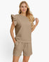 A woman modeling a matching beige lounge set consisting of a sleeveless top with ruffle details on the shoulders and matching drawstring shorts, set against a plain white studio background.
