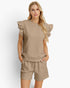 A woman modeling a matching beige lounge set featuring a textured short-sleeve top with ruffled shoulders and matching drawstring shorts, set against a plain light gray background.
