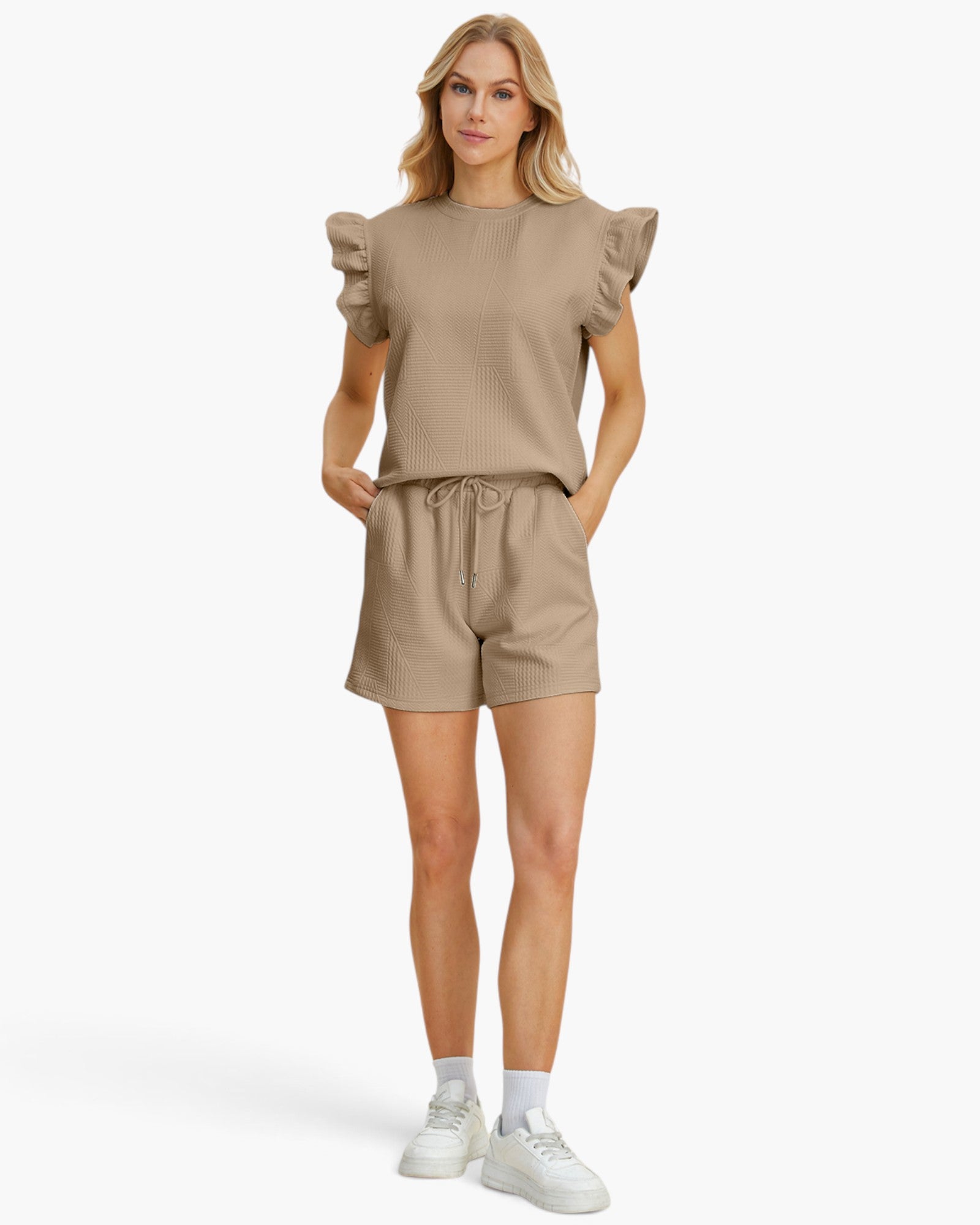 A woman modeling a matching beige lounge set consisting of a short-sleeve top with ruffled shoulders and matching drawstring shorts, paired with white socks and sneakers, against a plain white studio background.