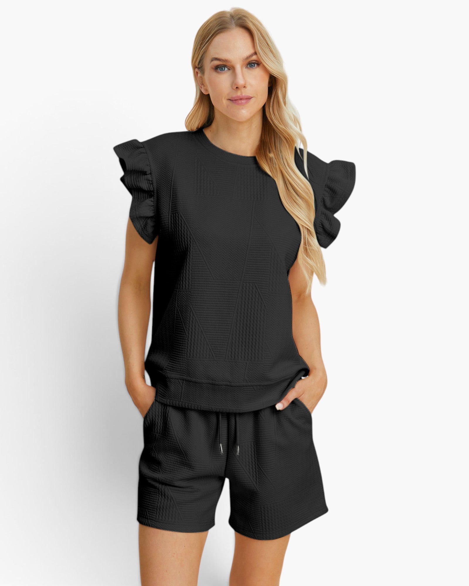 A woman modeling a black ribbed two-piece lounge set featuring a short-sleeve top with dramatic ruffled shoulders and matching drawstring shorts, set against a plain light gray studio background.