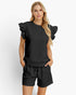A woman modeling a black ribbed two-piece lounge set featuring a short-sleeve top with dramatic ruffled shoulders and matching drawstring shorts, set against a plain light gray studio background.