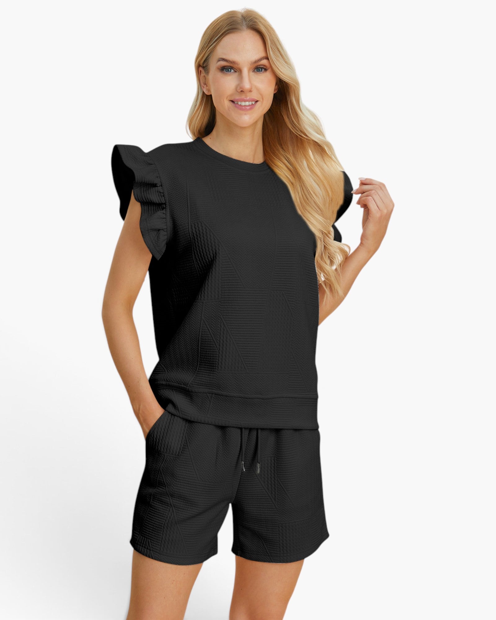 A woman modeling a black ribbed lounge set consisting of a sleeveless top with ruffled shoulder details and matching drawstring shorts, against a plain white studio background.
