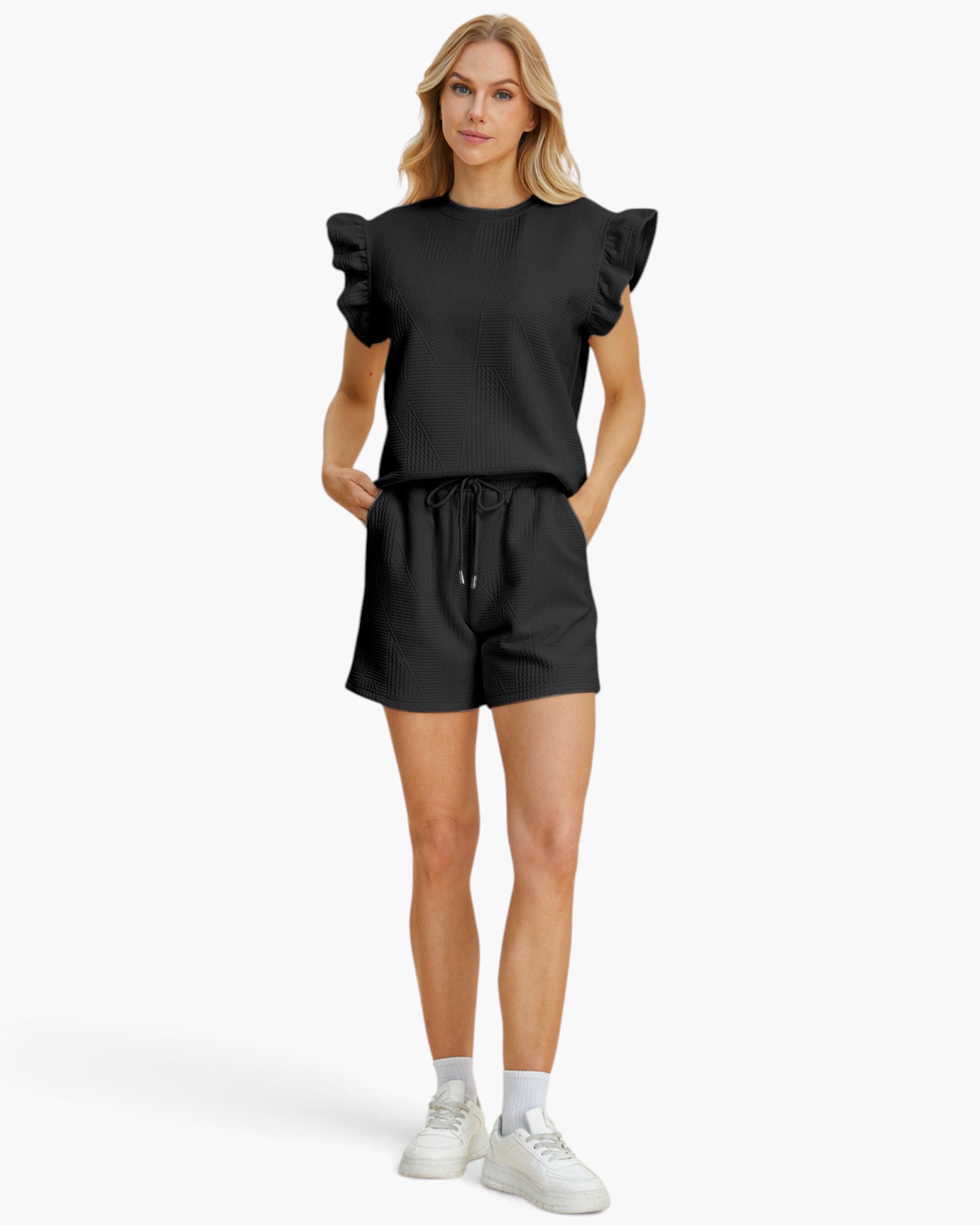 A woman modeling a black short-sleeve top with ruffled shoulders and matching black shorts, paired with white socks and white sneakers, against a plain light gray studio background.
