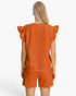 A woman shown from the back wearing a matching burnt orange textured knit set consisting of a short-sleeve top with ruffled shoulders and matching shorts, against a plain white studio background.