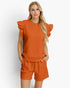 A woman modeling a matching burnt orange two-piece lounge set featuring a textured top with ruffled sleeves and coordinating shorts, set against a plain light gray background.