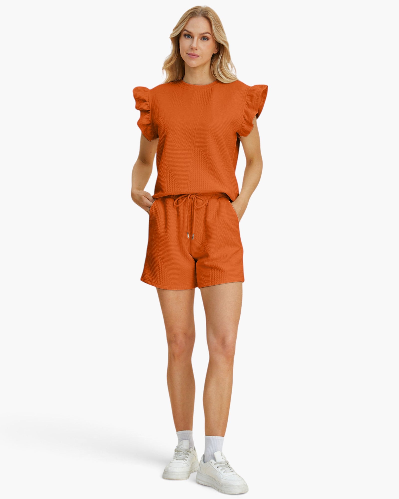 A woman modeling a matching burnt orange two-piece set consisting of a short-sleeve top with ruffled shoulders and matching drawstring shorts, paired with white sneakers and socks, against a plain white studio background.