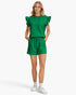 A woman modeling a bright green two-piece set consisting of a short-sleeve top with ruffled shoulders and matching drawstring shorts, paired with white socks and sneakers, against a plain white studio background.