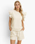 A woman modeling a cream-colored two-piece lounge set consisting of a textured short-sleeve top with ruffled shoulders and matching drawstring shorts, set against a plain light gray studio background.
