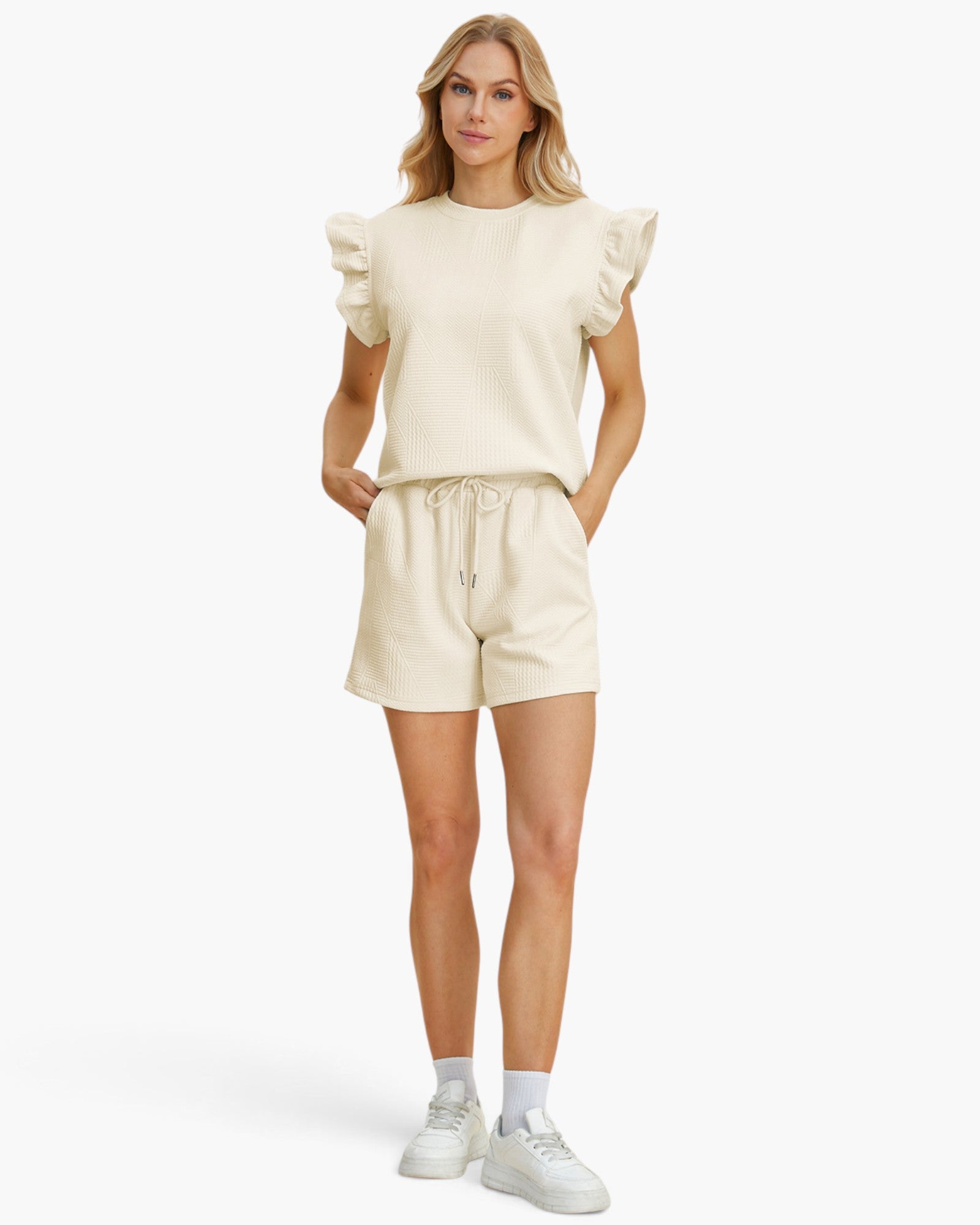 A woman modeling a matching cream-colored lounge set consisting of a short-sleeve top with ruffled shoulders and drawstring shorts, paired with white sneakers and socks, against a plain white studio background.