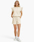 A woman modeling a matching cream-colored lounge set consisting of a short-sleeve top with ruffled shoulders and drawstring shorts, paired with white sneakers and socks, against a plain white studio background.