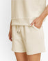 A close-up of a person wearing cream-colored textured shorts with a drawstring waist and side pockets, paired with a matching top against a plain light background.