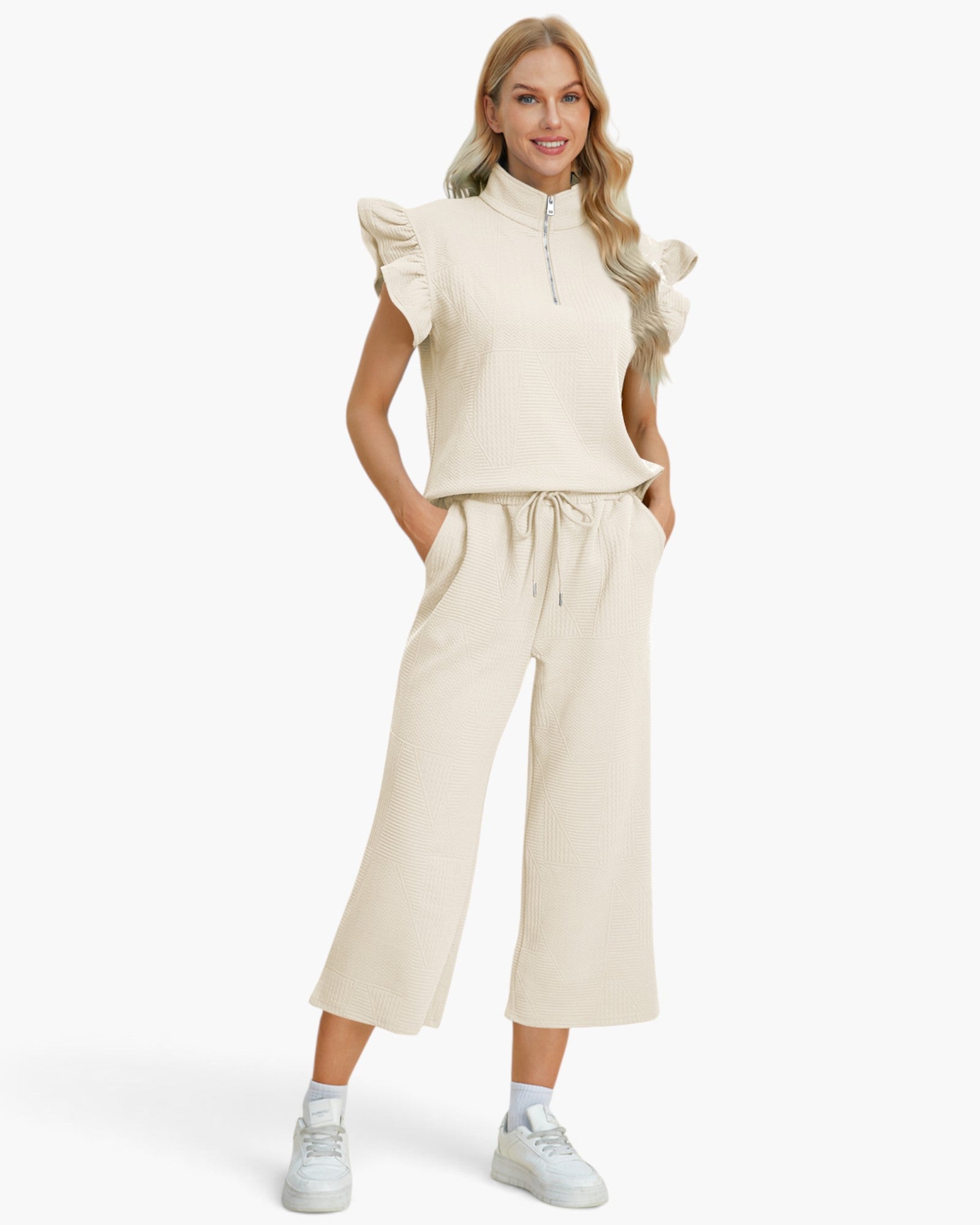 A woman modeling a matching cream-colored two-piece set consisting of a sleeveless ruffled mock-neck top with a half-zip and wide-leg cropped pants with a drawstring waist, paired with white sneakers against a light gray studio background.