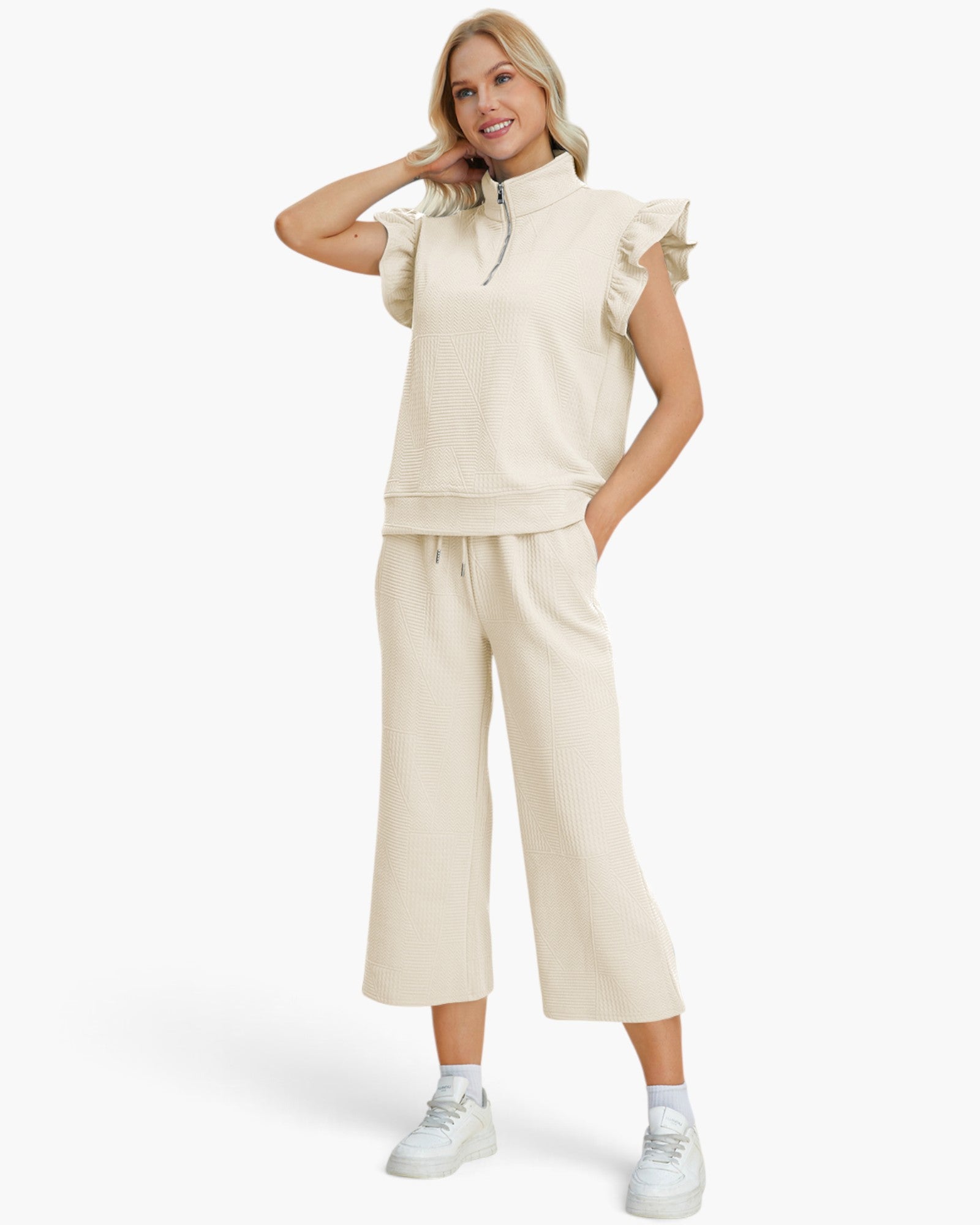 A woman modeling a matching cream-colored loungewear set consisting of a short-sleeve ruffled mock-neck zip sweatshirt and wide-leg cropped sweatpants, paired with white sneakers against a plain white background.