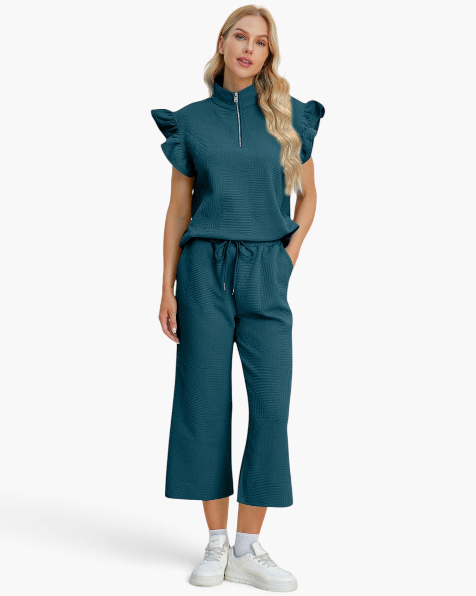A woman modeling a matching teal two-piece outfit consisting of a short-sleeve top with ruffled shoulders and a half-zip neckline, paired with wide-leg cropped pants with a drawstring waist, on a light gray studio background.