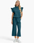 A woman modeling a matching teal two-piece outfit consisting of a short-sleeve top with ruffled shoulders and a half-zip neckline, paired with wide-leg cropped pants with a drawstring waist, on a light gray studio background.
