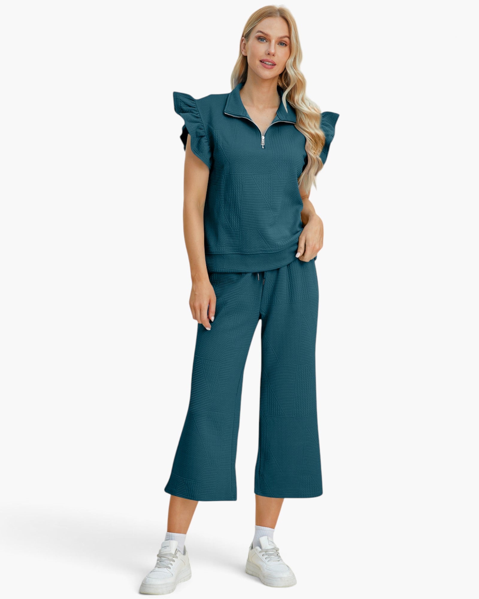 A woman modeling a matching teal two-piece lounge set consisting of a ruffle-sleeve half-zip top and wide-leg cropped pants, paired with white sneakers against a light gray studio background.