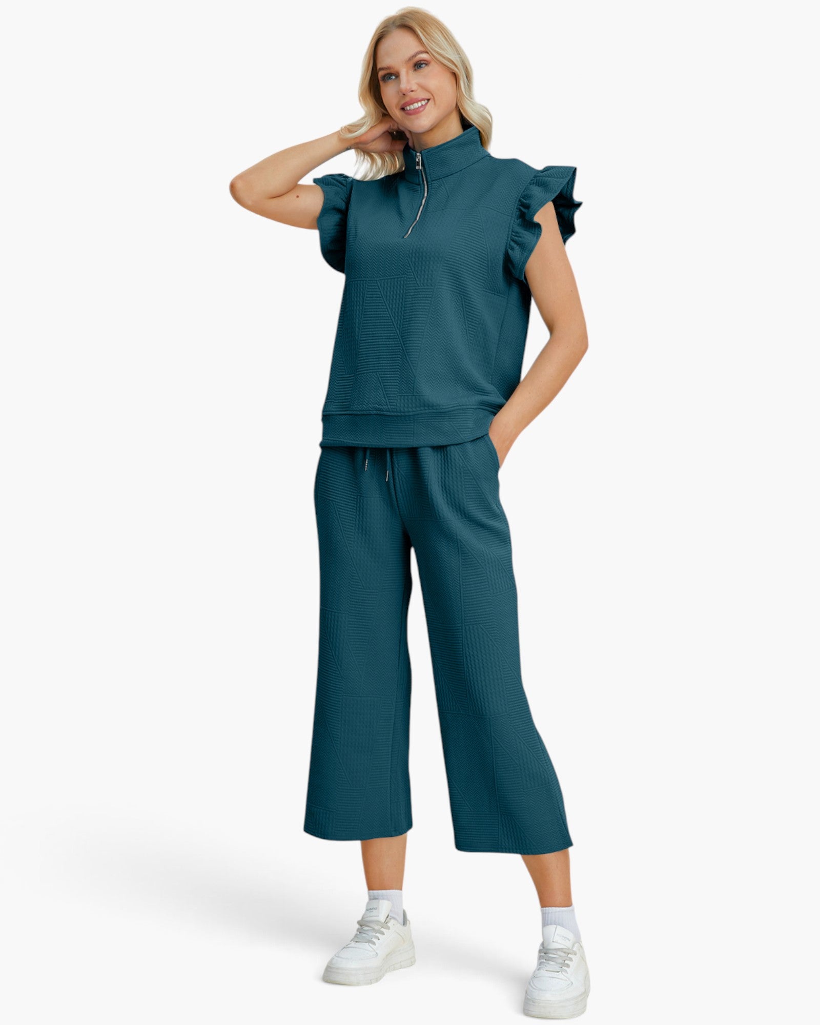 A woman modeling a matching two-piece tracksuit in a deep teal color, featuring a ruffle-trimmed short-sleeve top with a half-zip and matching wide-leg cropped pants, paired with white sneakers against a light gray background.
