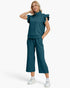 A woman modeling a matching two-piece tracksuit in a deep teal color, featuring a ruffle-trimmed short-sleeve top with a half-zip and matching wide-leg cropped pants, paired with white sneakers against a light gray background.