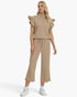 A woman modeling a matching beige two-piece set consisting of a short-sleeve top with ruffled shoulders and a half-zip neckline, paired with wide-leg cropped pants with a drawstring waist, on a light gray studio background.
