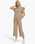 A woman modeling a matching beige lounge set consisting of a short-sleeve ruffle-shoulder quarter-zip top and wide-leg cropped pants, paired with white sneakers against a light gray studio background.