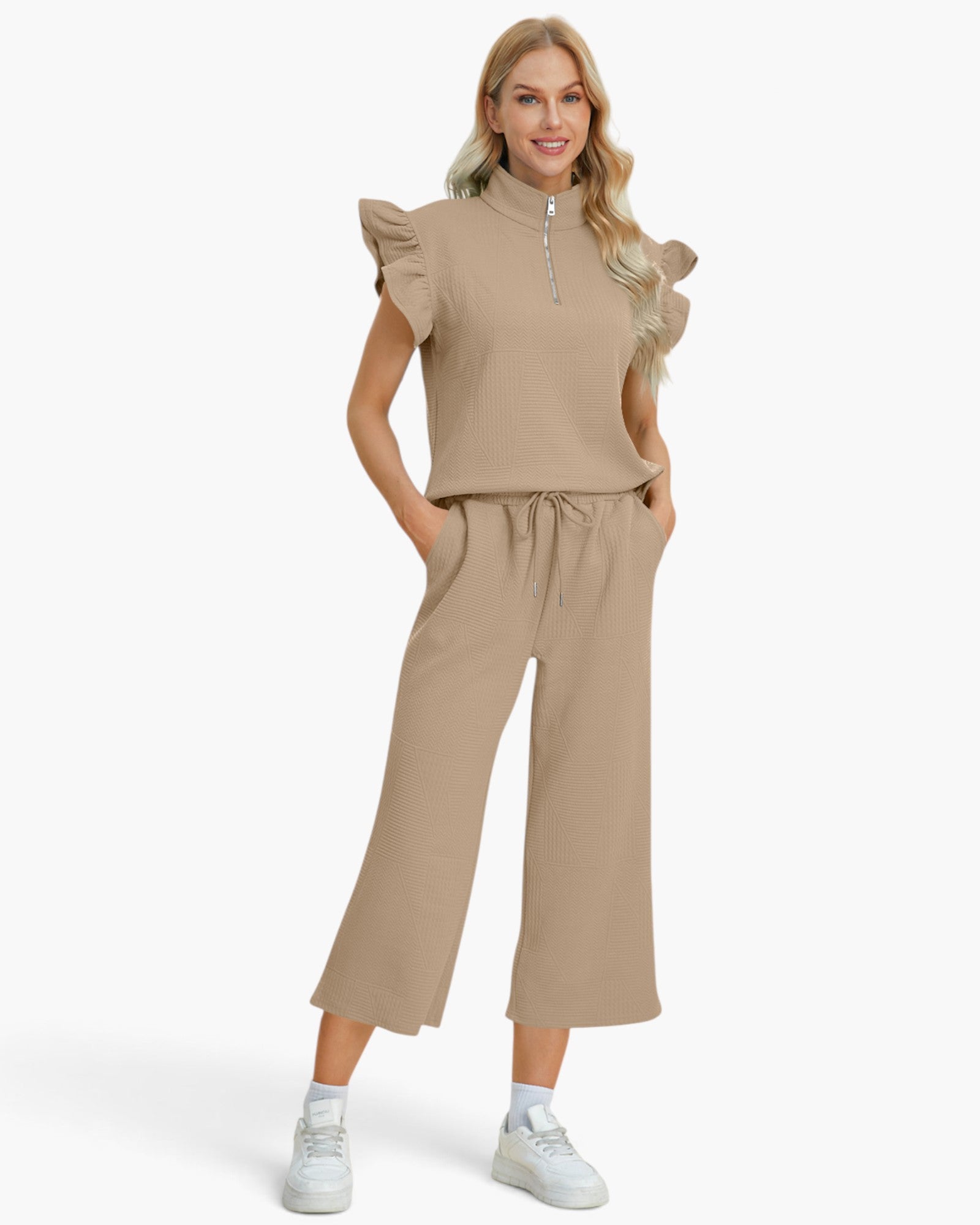 A woman modeling a matching beige two-piece set consisting of a sleeveless ruffled mock-neck top with a short silver zipper and matching wide-leg cropped pants with a drawstring waist, paired with white sneakers against a light gray studio background.