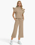 A woman modeling a matching beige two-piece set consisting of a sleeveless ruffled mock-neck top with a short silver zipper and matching wide-leg cropped pants with a drawstring waist, paired with white sneakers against a light gray studio background.